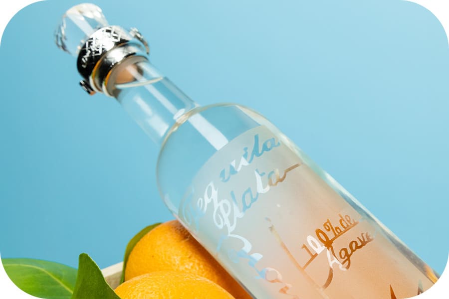 Botella de Tequila Don Ramón Plata, 100% agave, acompañada de limones sobre fondo azul.