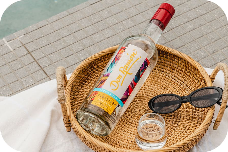 Botella de Tequila Don Ramón en canasta con gafas de sol sobre una mesa con mantel blanco, detras se ve un piso cuadriculado.